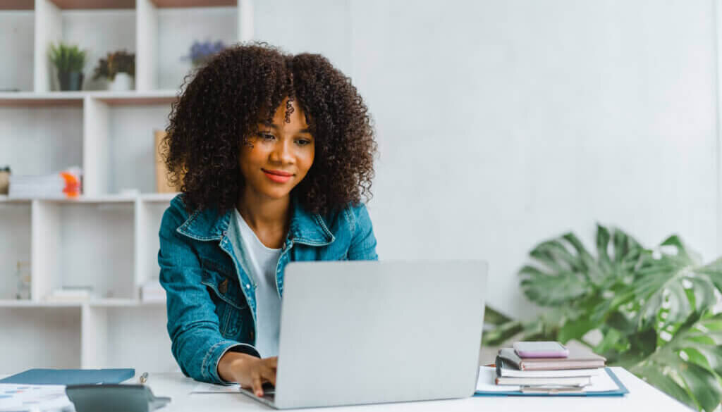 Woman working on laptop at desk with books and calculator. A plant and stacked square shelving is in the background.