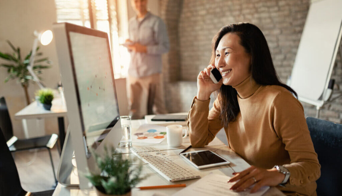 Woman smiling while on phone and looking at computer screen while sitting at her desk.