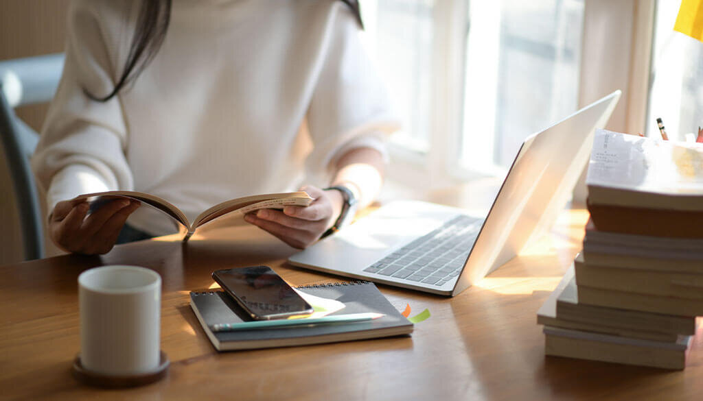 Person holding an open book at desk with laptop, phone, and books surrounding her.