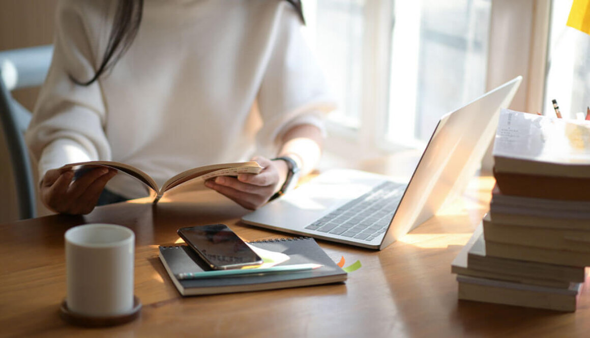Person holding an open book at desk with laptop, phone, and books surrounding her.