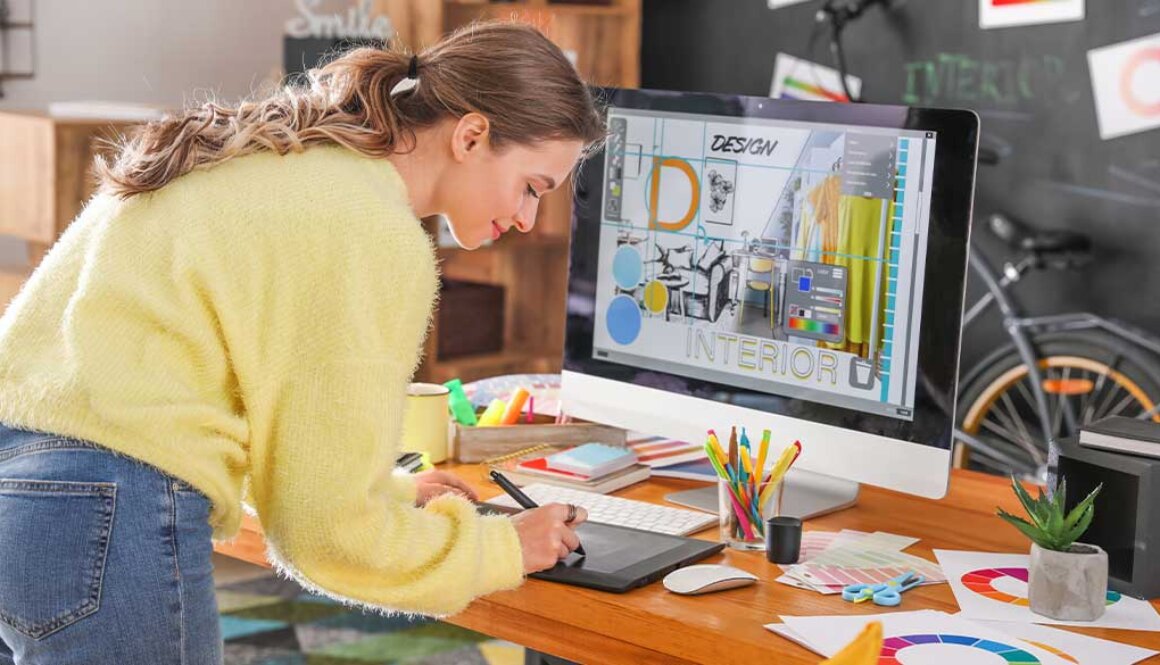 Young woman working at a design desk with computer while writing on a tablet.