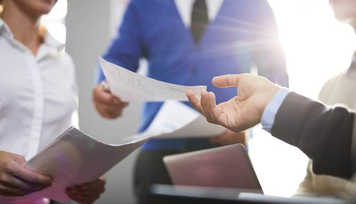 A blurry background of business team holding papers and working on laptop and a hand reaching out grabbing a paper in the foreground.