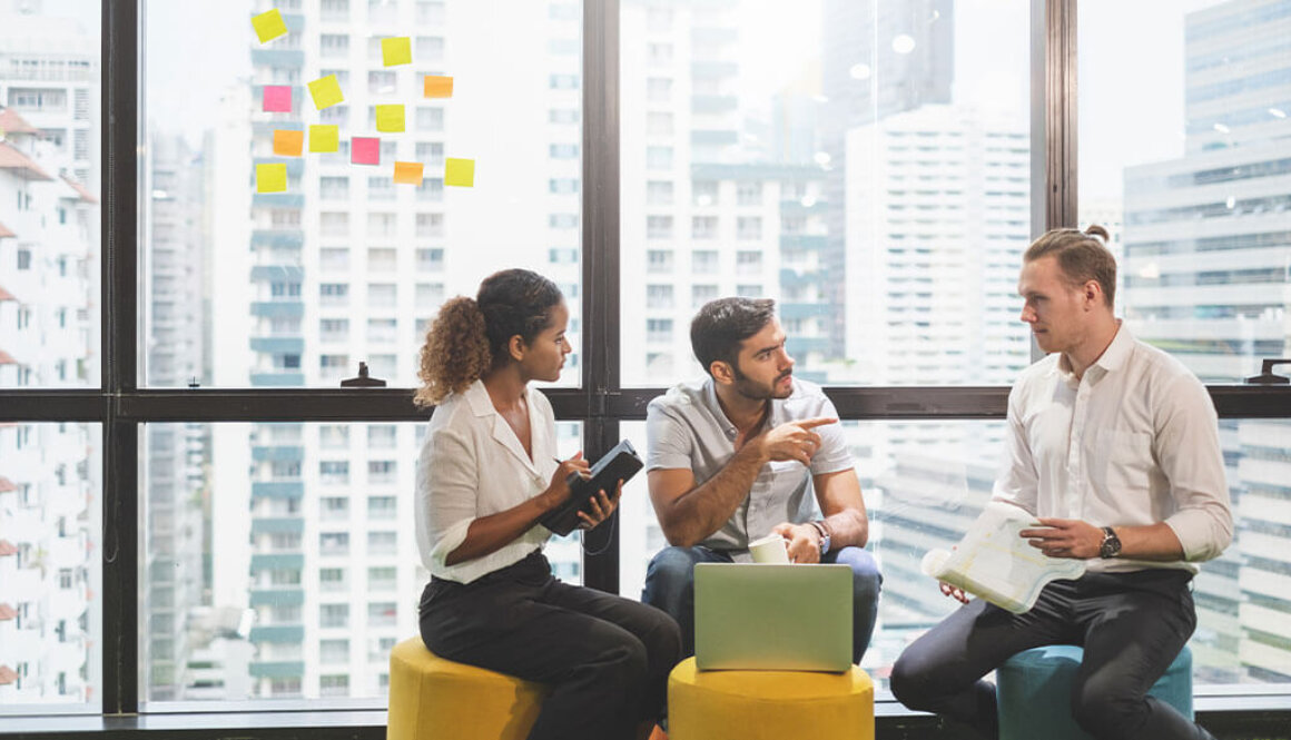 Team of three people sitting on cushion stools with sticky notes posted on a window in the background with city buildings.