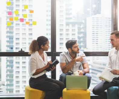 Team of three people sitting on cushion stools with sticky notes posted on a window in the background with city buildings.