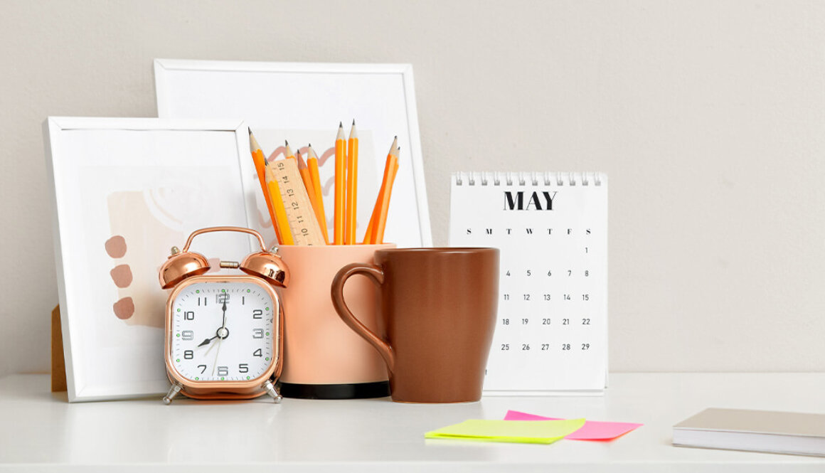An organized desk photo with photo frames, brown mug, modern retro alarm clock, pencils in a beige cup holder, a calendar, sticky notes, and a notebook.