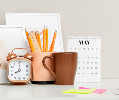 An organized desk photo with photo frames, brown mug, modern retro alarm clock, pencils in a beige cup holder, a calendar, sticky notes, and a notebook.