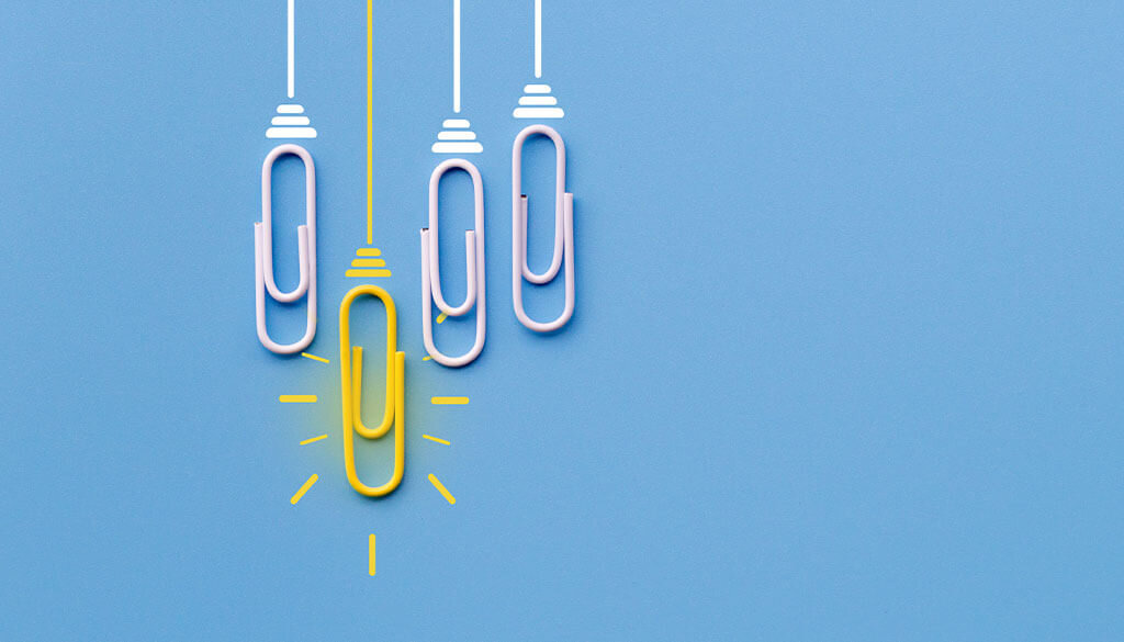 Four paperclips laying on blue background while one bright yellow paperclip hangs lower.
