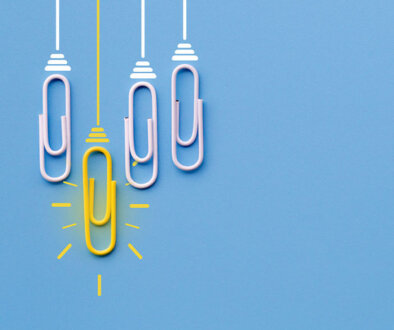 Four paperclips laying on blue background while one bright yellow paperclip hangs lower.